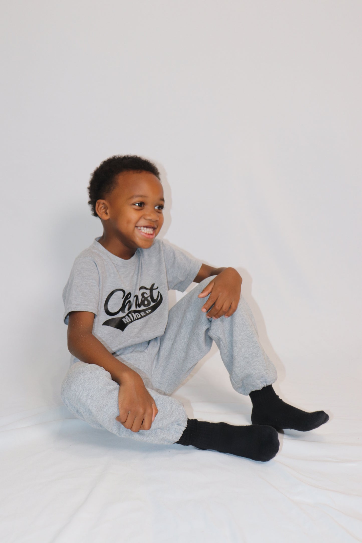 Boy sitting and smiling in a gray Christ Minded shirt with matching joggers and black socks