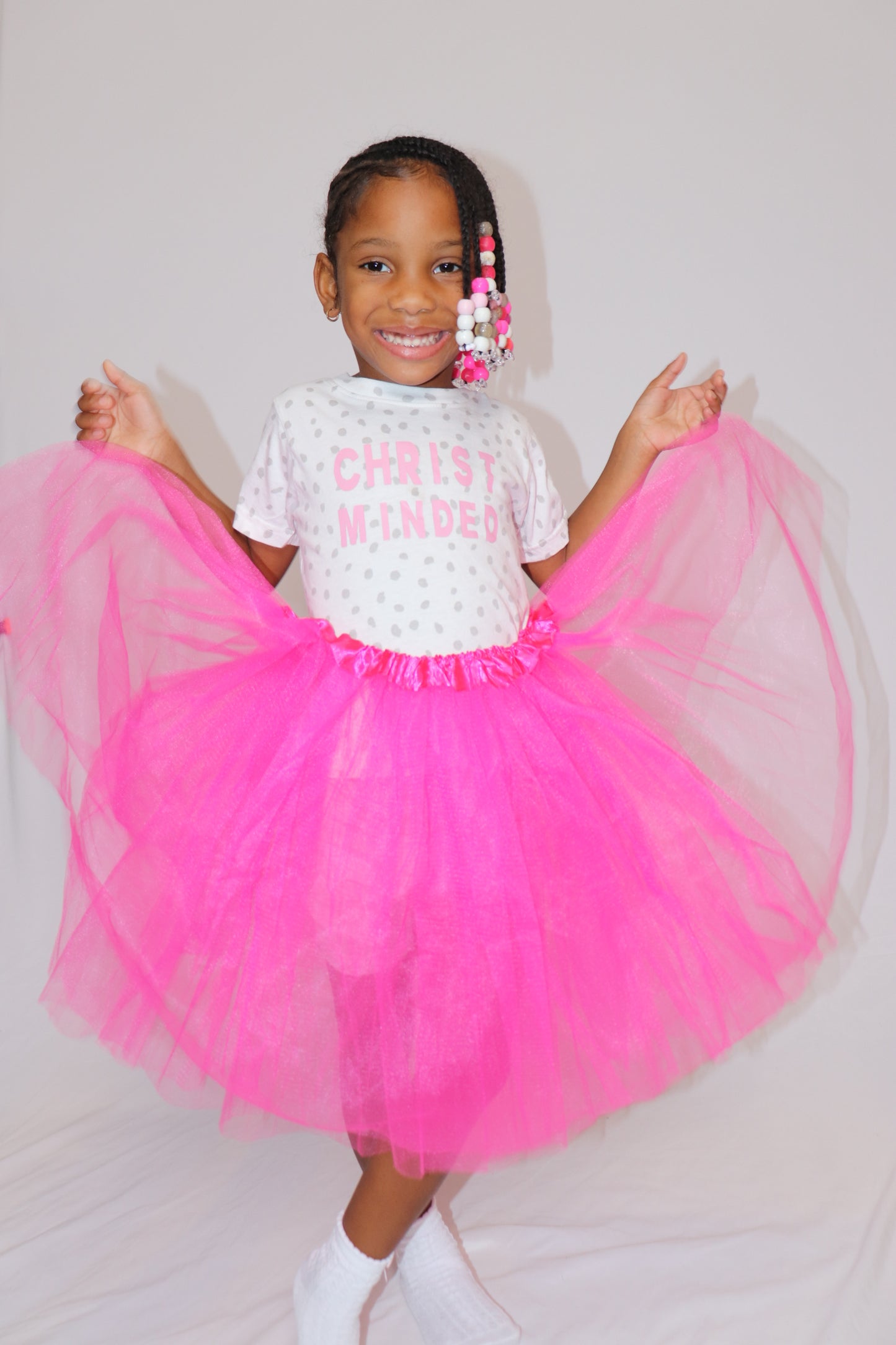 Young girl wearing a Christ Minded polka dot t-shirt with a bright pink tutu, smiling joyfully in a faith-based kids apparel photoshoot