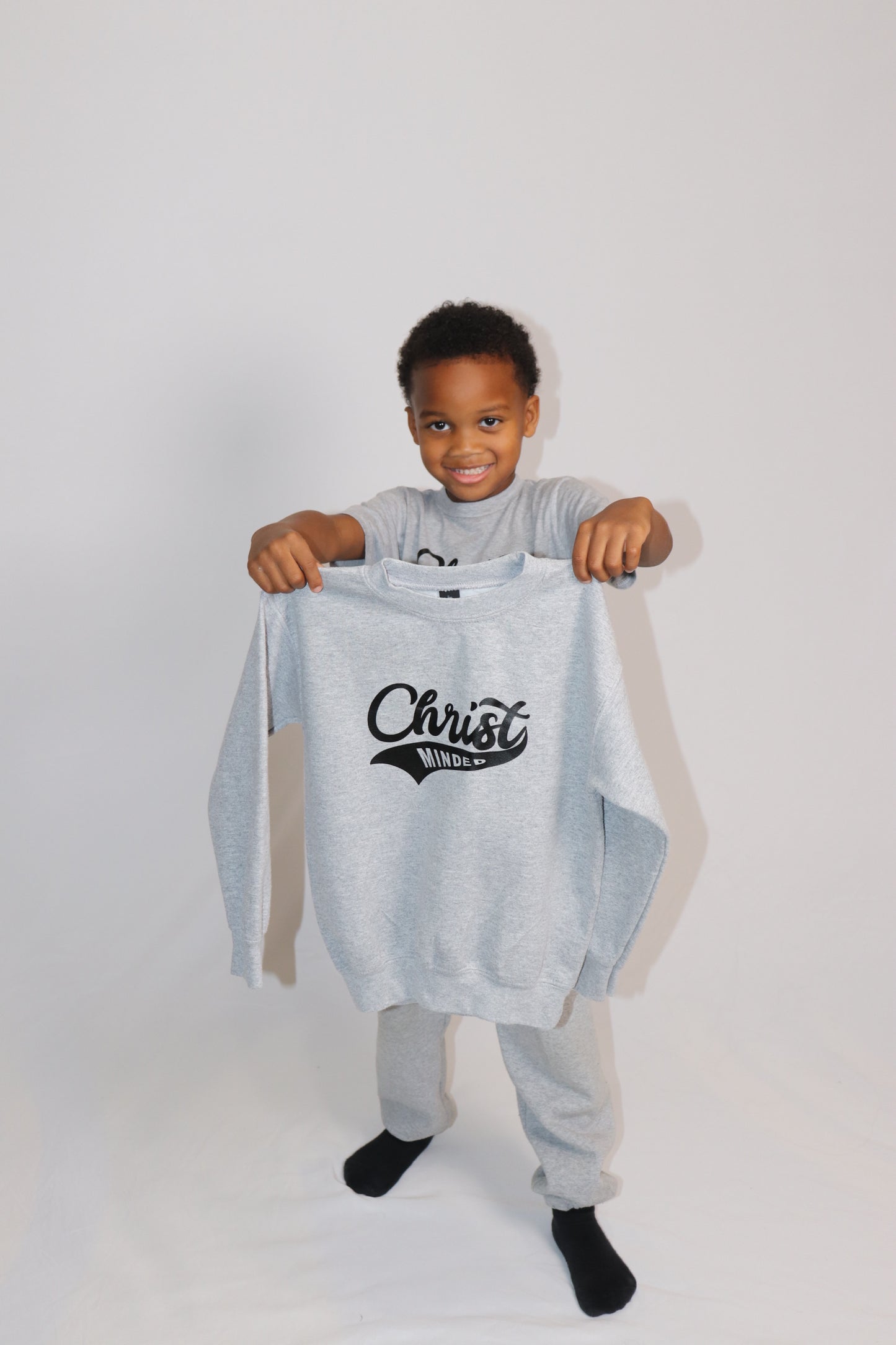 Smiling boy holding up a gray Christ Minded sweatshirt while wearing matching sweatpants