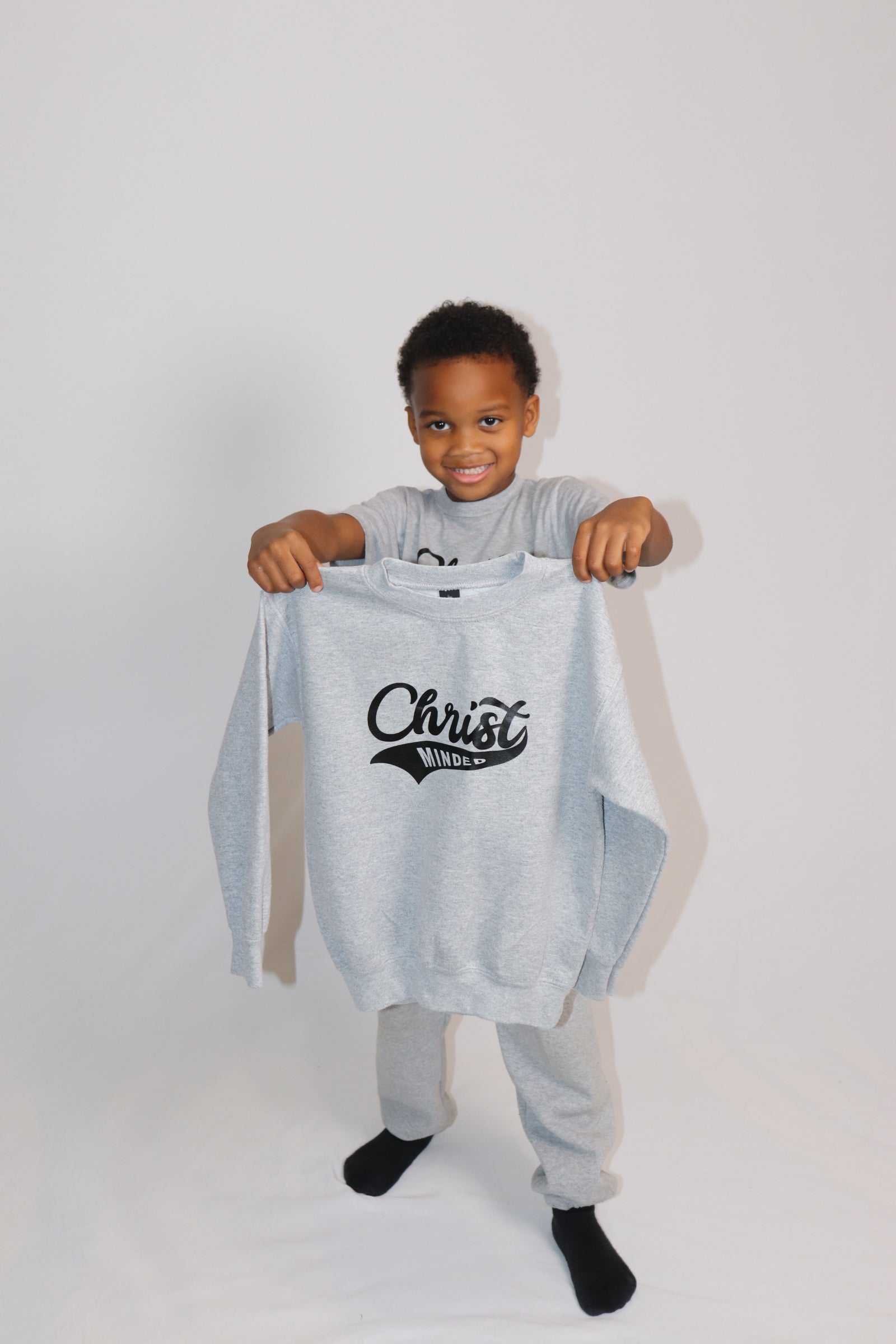 Smiling boy holding up a gray Christ Minded sweatshirt while wearing matching sweatpants